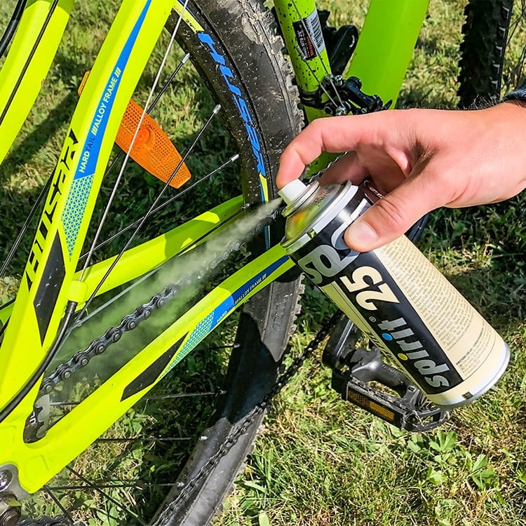Zestaw do konserwacji roweru Spirit, składający się z czterech sprayów, używany do smarowania łańcucha. Na zdjęciu widać rękę trzymającą spray, z którego wydobywa się mgła, aplikująca preparat na łańcuch rowerowy. Tło to trawa.