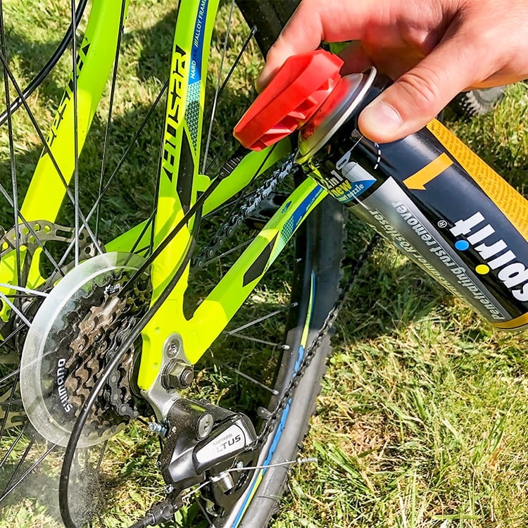 Zestaw do konserwacji roweru Spirit, składający się z dwóch sprayów. Ujęcie z boku, ręka trzymająca spray skierowany na łańcuch i zębatkę roweru. Rower w intensywnym zielonym kolorze, z metalicznymi elementami. Tło naturalne, trawiaste.