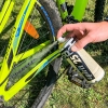 Zestaw do konserwacji roweru Spirit zawiera dwa spray’e. Na zdjęciu widoczna jest ręka aplikująca spray na łańcuch roweru. Rower ma jasną, zieloną ramę z niebieskimi i czarnymi detalami, a tło to trawa.
