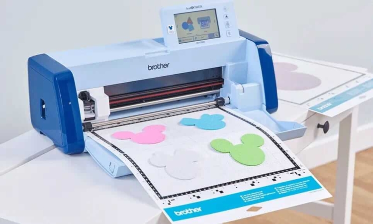 Ploter tnący Brother ScanNCut SDX2250 w kolorze niebieskim z wyświetlaczem, z rolką materiału na stole, z wyciętymi kształtami w kolorze różowym, niebieskim, zielonym i białym, w aranżacji domowej. Widoczna podstawa z oznaczeniem.