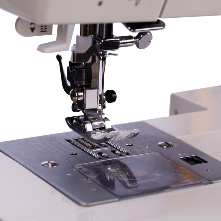 Maszyna do szycia Minerva MC110 PRO, widok z bliska na stopkę oraz mechanizm przesuwu tkaniny. Widać szereg otworów do szycia oraz przezroczystą osłonę. Elementy metalowe i plastikowe w kolorze białym i srebrnym.