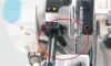 Zbliżenie na mechanizm owerloka Jack C5F-4, ukazujące elementy sterujące, w tym dwa otwory oraz przycisk w kolorze niebieskim. Widoczne są także metalowe mocowania oraz przewód, co wskazuje na zaawansowaną konstrukcję maszyny.