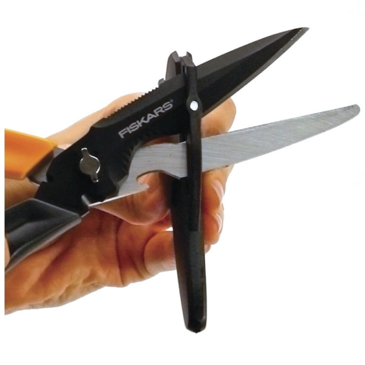Nożyczki wielofunkcyjne Fiskars Cuts+More o długości 23 cm, z czarnymi ostrzami i ergonomiczną rączką. Ujęcie z boku, w dłoni, z widoczną funkcją cięcia. Ostrza wykonane ze stali, z dodatkowymi elementami do otwierania oraz śrubokrętem.