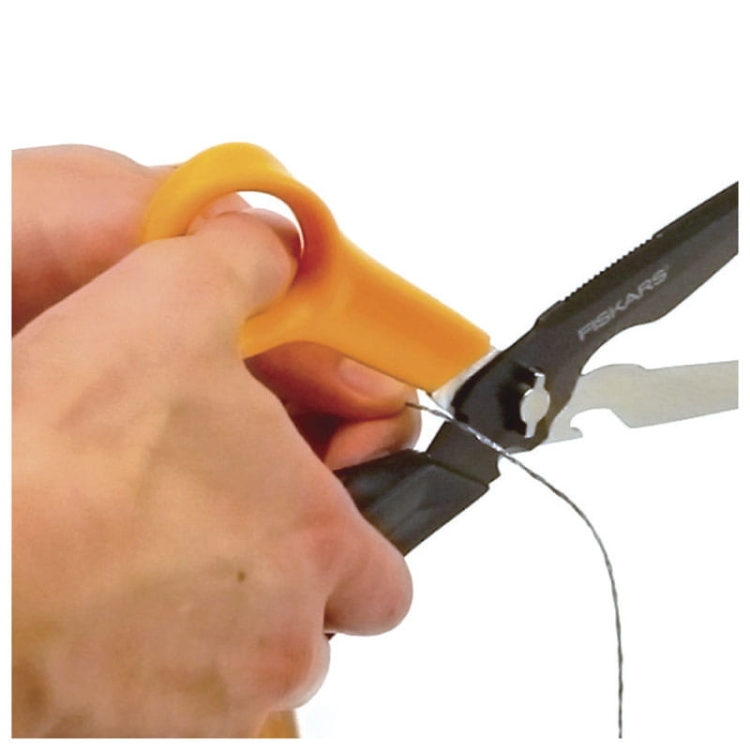 Nożyczki wielofunkcyjne Fiskars Cuts+More 1000809 mają żółty uchwyt z ergonomiczną formą i czarne ostrze. Na zdjęciu ukazano rękę trzymającą nożyczki, które tną cienki drut. Ujęcie z bliska, skupione na detalu użycia.