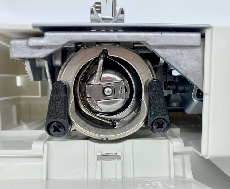 Zbliżenie na mechanizm bębenka maszyny do szycia Minerva Next 532A. Widoczna metalowa część bębna z czarnymi uchwytami po bokach. Tło w jasnym kolorze, co podkreśla detale mechanizmu.