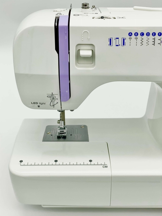 Maszyna do szycia Minerva Next 141D II w kolorze białym z fioletowymi detalami. Widoczny stół z miarką, metalowa stopka i panel z wyborem st stitchów. LED-owe oświetlenie, mechanizm nawijania nici oraz przyciski funkcyjne. Ujęcie z boku.