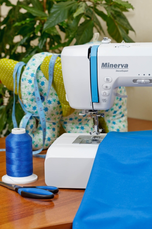 Maszyna do szycia Minerva DecorExpert w kolorze białym z niebieskimi detalami, umieszczona na drewnianym stole. Obok niebieska szpula nici, nożyczki oraz rozłożony niebieski materiał. W tle widoczne kolorowe tkaniny i miara krawiecka.