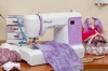 Maszyna do szycia Minerva DecorMaster w białej i fioletowej obudowie, z tkaniną w odcieniach fioletu pod stopką. Widoczne przyciski funkcyjne oraz wyświetlacz. Obok leżą nożyczki i szpilki w poduszce, a w tle kolorowe pudełka z motywem kwiatowym.
