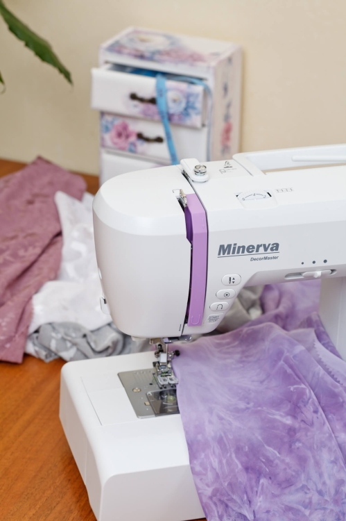 Maszyna do szycia Minerva DecorMaster w kolorze białym z fioletowymi akcentami, umieszczona na drewnianym blacie. Obok widoczne materiały w odcieniach fioletu i szarości, w tle ozdobne pudełko z szufladkami. Zbliżenie na igłę i stopkę maszyny.