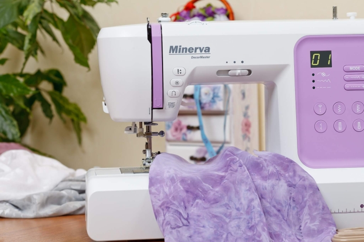 Maszyna do szycia Minerva DecorMaster w kolorze białym z fioletowymi akcentami, z widocznym wyświetlaczem i przyciskami. Z przodu wystaje materiał w odcieniu lawendowym. Ujęcie z boku, na tle roślinności, prezentuje detale maszyny i szytego materiału.