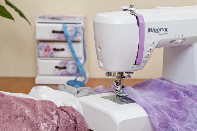 Maszyna do szycia Minerva DecorMaster w kolorze białym z fioletowymi akcentami, ustawiona z przodu. Na stole widoczne materiały w odcieniach różu i bieli oraz miara krawiecka. Tło to mebel z szufladami ozdobionymi kwiatowym wzorem.