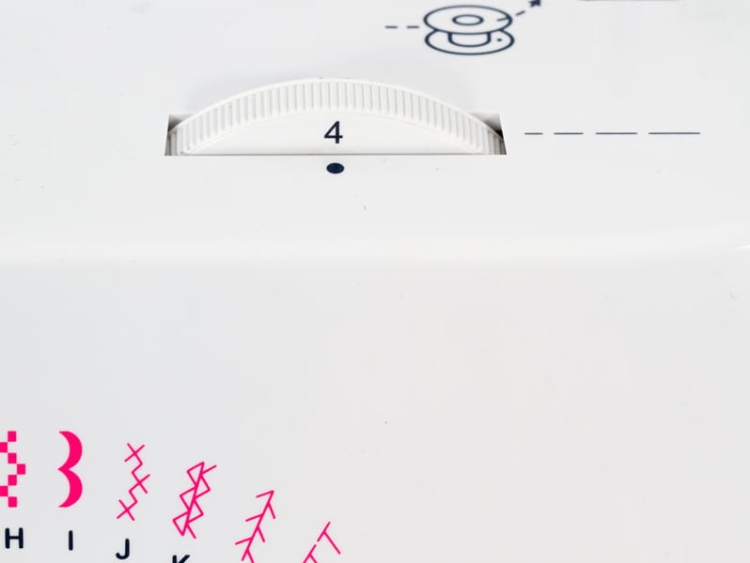 Biały panel maszyny do szycia JUKI HZL-353 ZR-A z pokrętłem ustawienia o numerze 4, obok ilustracja szpulki. Widoczne kolorowe napisy i wzory szwów. Ujęcie z bliska na górną część maszyny.