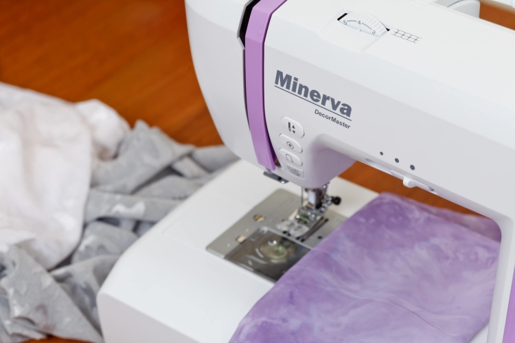 Maszyna do szycia Minerva DecorMaster w kolorze białym z różowymi akcentami, umieszczona na stole. Widoczny jest uchwyt do szycia oraz materiał w odcieniu fioletowym, częściowo na stelażu. Tło w odcieniach drewna, tworzy naturalny kontekst użytkowania.