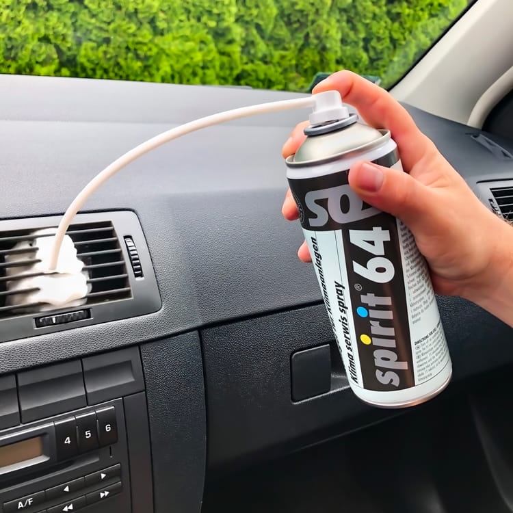 Środek do odgrzybiania klimatyzacji w sprayu 400 ml Spirit 64 trzymany w dłoni, z elastyczną rurką skierowaną w stronę kratki wentylacyjnej. Produkt znajduje się na desce rozdzielczej samochodu. Tło stanowi zielony żywopłot.