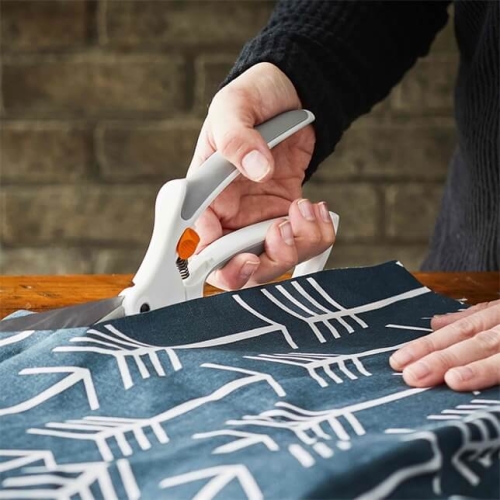 Nożyce krawieckie Fiskars Easy Action 1070070 o długości 26 cm, trzymane w prawej ręce, tną materiał w kolorze granatowym z białym wzorem. Przeznaczone do precyzyjnego cięcia, z ergonomiczną rączką i mechanizmem wspomagającym. Tło drewniane.