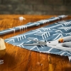 Nożyce krawieckie Fiskars Easy Action o długości 26 cm, z białymi uchwytami i pomarańczowym przyciskiem, leżą na niebieskim materiale z białym wzorem. W tle widoczne elementy krawieckie, takie jak taśma i szpuleczka, na drewnianym blacie.