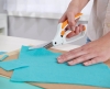 Nożyce krawieckie Fiskars Easy Action 1070070 o długości 26 cm, z białą rączką i pomarańczowymi akcentami, są używane do cięcia błękitnego filcu. Ujęcie z boku ukazuje rękę trzymającą nożyce nad materiałem, z widoczną powierzchnią roboczą.