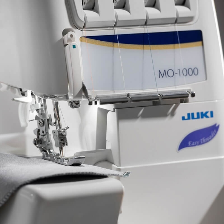 Owerlok JUKI MO-1000 z widoczną stopką i igłą, szyjący szary materiał. Maszyna ma cztery nitki, z niebieskim i złotym logo na obudowie. Ujęcie z boku, ukazujące detale mechanizmu i łatwe wprowadzanie nici.