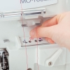 Owerlok JUKI MO-1000 w zbliżeniu, z ręką nawlekającą czerwoną nitkę przez otwór na górze maszyny. Widoczna instrukcja z symbolami na panelu. Biała obudowa urządzenia z metalowymi elementami.
