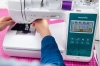 Maszyno-hafciarka Brother F580 w kolorze białym z zielonym panelem sterującym, z widocznym ekranem LCD. Użytkownik trzyma materiał pod stopką, a na górze widać szpulę z fioletową nitką. Kąt ujęcia z boku, na różowym tle.