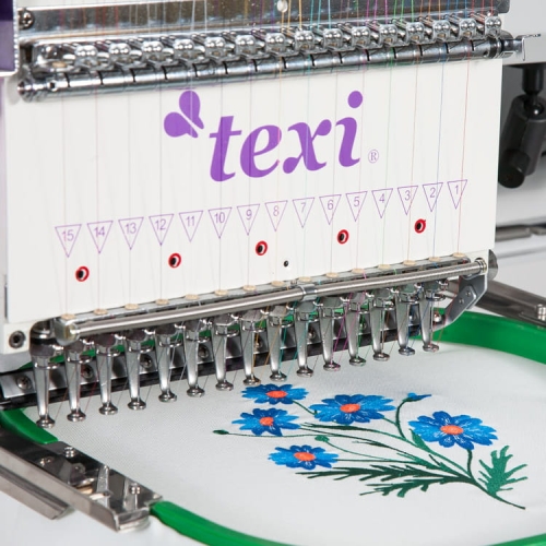 Hafciarka TEXI 1501 TS PREMIUM, 15-igłowa, z widocznym logo texi. Zbliżenie na igły i haft z niebieskimi kwiatami na białym tle, umieszczonym w zielonym tamborku. Maszyna w pozycji roboczej, gotowa do szycia.