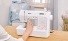 Maszyna do szycia Brother KD40S Little Angel w białej obudowie, z wyświetlaczem LCD. Widoczna ręka użytkownika ustawiająca program szycia. W tle szara bluza z tęczą. Na maszynie kolorowe wzory i logo Brother. Ułożona na drewnianym blacie.