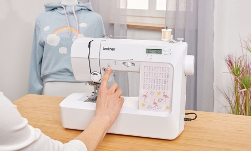 Maszyna do szycia Brother KD40S Little Angel w białej obudowie, z wyświetlaczem LCD. Widoczna ręka użytkownika ustawiająca program szycia. W tle szara bluza z tęczą. Na maszynie kolorowe wzory i logo Brother. Ułożona na drewnianym blacie.