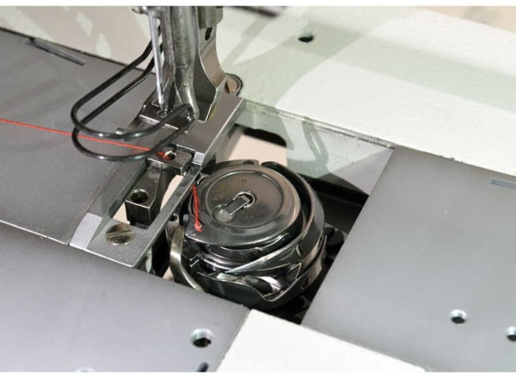 Zbliżenie na mechanizm lamowarki KRAFFT KF-1245V, pokazujący igłę, prowadzenie nitki oraz szpulę z nicią. Widoczny metalowy element z otworem oraz potrójny transport. Tło w jasnym kolorze, ułatwiające identyfikację detali.