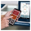 Maszyno-hafciarka Husqvarna Viking DESIGNER SAPPHIRE 85, zbliżenie na rękę trzymającą smartfon z aplikacją do obsługi maszyny. Na ekranie widoczny kolorowy wzór haftu. Tło maszyny z elementami sterującymi w kolorze niebieskim.
