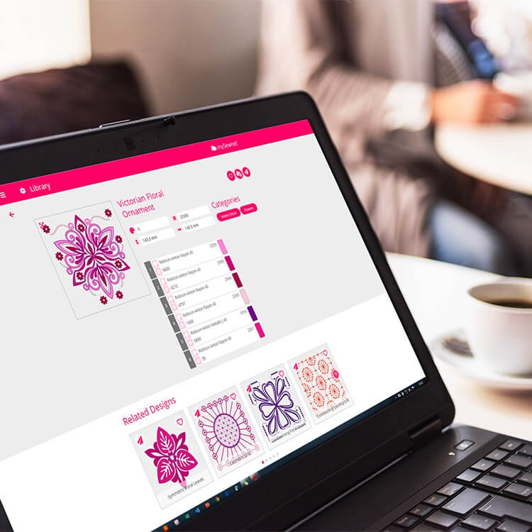 Ekran laptopa z interfejsem maszyno-hafciarki Husqvarna Viking DESIGNER EPIC 2, wyświetlający projekt Victorian Floral Ornament oraz powiązane wzory. Tło rozmyte, widać filiżankę z napojem.