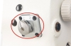 Zbliżenie na metalowy mechanizm w maszynie do szycia Zygzak JACK JK-T2284B-4E. Widoczne elementy: okrągły przycisk z wcięciem i śrubą, otoczony czterema śrubami mocującymi, na białym tle obudowy maszyny.