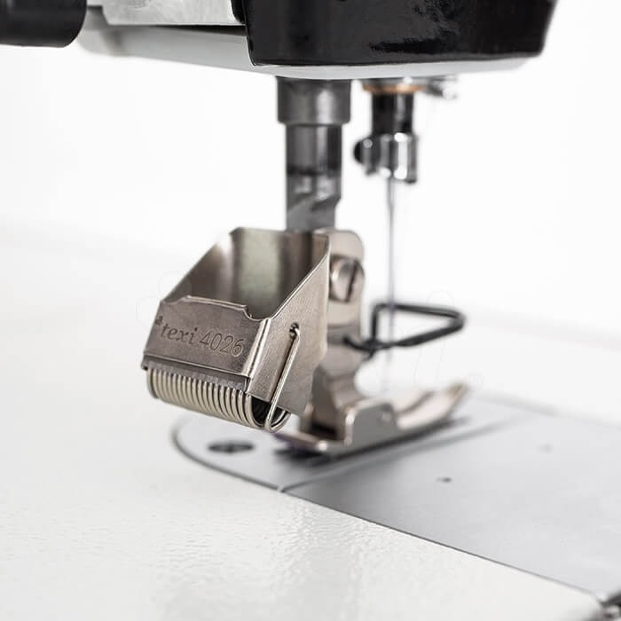 Obcinacz nici TEXI 4026 zamontowany na maszynie przemysłowej, wykonany z metalu, z widocznym napisem TEXI 4026. Produkt w pozycji roboczej, z bliska ukazuje mechanizm obcinający oraz elementy mocujące. Tło proste, jasne.