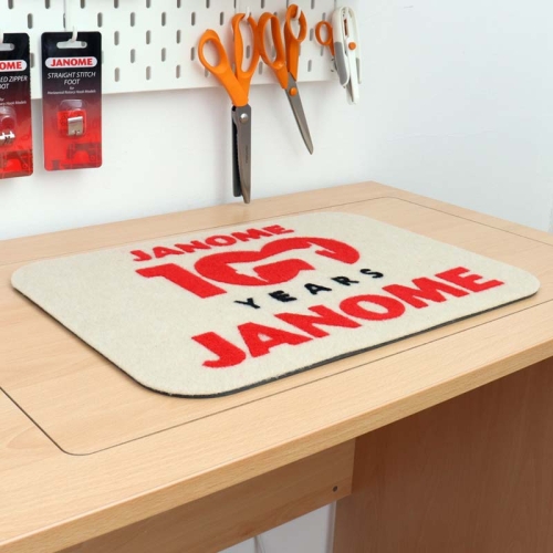 Podkładka filcowa Janome w kolorze beżowym z czerwonym napisem JANOME 100 YEARS i czarnym YEARS. Wymiary 44,5 x 32,5 cm, leżąca na drewnianym blacie, w otoczeniu narzędzi krawieckich.