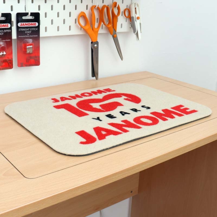 Podkładka filcowa Janome w kolorze beżowym z czerwonym napisem JANOME 100 YEARS i czarnym YEARS. Wymiary 44,5 x 32,5 cm, leżąca na drewnianym blacie, w otoczeniu narzędzi krawieckich.