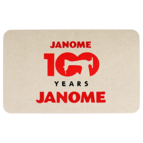 Podkładka filcowa Janome w kolorze beżowym, o wymiarach 68,5 x 42,5 cm. Na powierzchni widoczne logo Janome oraz napis 100 YEARS w czerwonym i czarnym kolorze. Produkt leży na płasko, bez dodatkowych elementów w tle.
