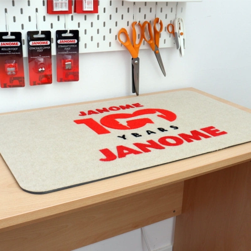 Podkładka filcowa Janome w kolorze beżowym, o wymiarach 68,5 x 42,5 cm, z czerwonym logo JANOME 100 YEARS. Ułożona na blacie roboczym, w otoczeniu narzędzi krawieckich, takich jak nożyczki i akcesoria.