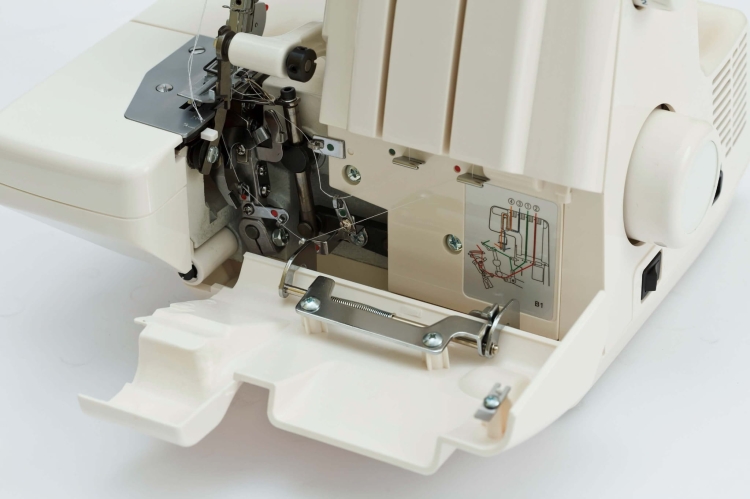 Owerlok Minerva M740DS 4-nitkowy z widocznym otwartym mechanizmem, metalowymi elementami i instrukcją montażu na obudowie. Kolor obudowy jasny, z dodatkiem srebrnych akcentów. Widoczny uchwyt do podawania materiału.