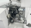 Widok wnętrza owerloka Minerva ML3314, ukazujący mechanizm z metalowymi elementami, sprężynami oraz prowadnikami nici. Widziane są oznaczenia kolorystyczne oraz pokrętło do regulacji. Maszyna jest w pozycji otwartej, co eksponuje jej detale konstrukcyjne