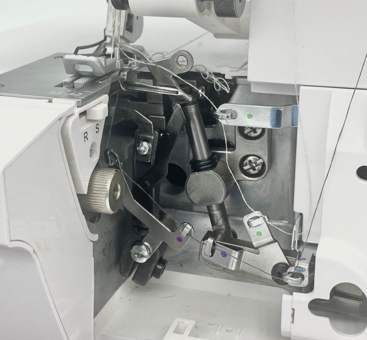 Widok wnętrza owerloka Minerva ML3314, ukazujący mechanizm z metalowymi elementami, sprężynami oraz prowadnikami nici. Widziane są oznaczenia kolorystyczne oraz pokrętło do regulacji. Maszyna jest w pozycji otwartej, co eksponuje jej detale konstrukcyjne
