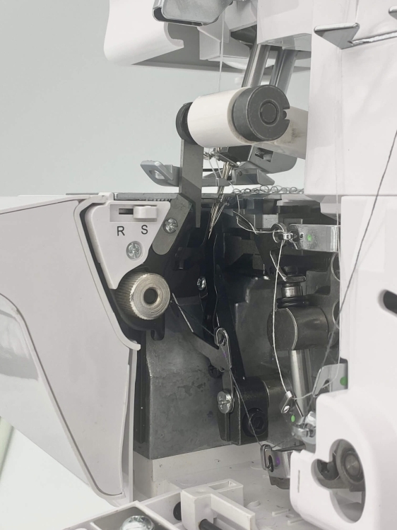 Owerlok Minerva ML3314, widok z boku na wnętrze maszyny. Widoczne elementy mechanizmu, w tym rolka z nicią, metalowe części oraz oznaczenia R i S. Obudowa w kolorze białym, z częściami w metalicznym odcieniu.