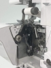 Owerlok Minerva ML3314 z widocznym mechanizmem, w tym rolką z białą nicią, metalowymi elementami i dźwigniami. Ujęcie z boku, ukazujące wnętrze maszyny do szycia, na jasnym tle.