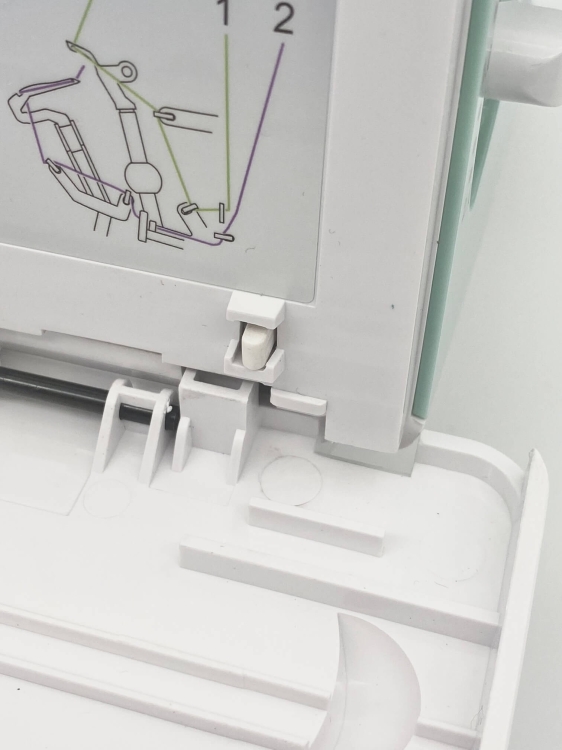 Zbliżenie na elementy mechaniczne owerloka Minerva ML3314. Widoczna instrukcja w formie ilustracji z numerami, pokazująca sposób nawlekania nitek. Białe przyciski i plastikowa obudowa w kolorze białym oraz delikatna zielona część.
