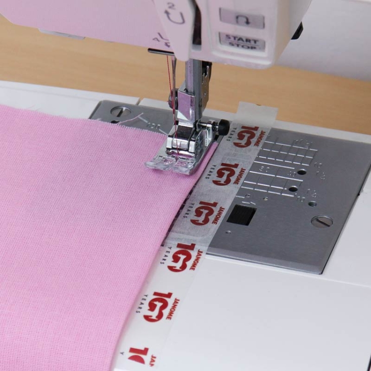 Taśma do szycia krawędziowego Janome 15 mm x 3 m w białym kolorze, z nadrukiem w czerwonym logo Janome. Widoczna na maszynie do szycia, używana do wykończenia różowego materiału. Kąt ujęcia z góry, z bliska na detal szycia.