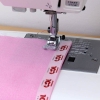 Taśma do szycia krawędziowego Janome 15 mm x 3 m w kolorze białym z czerwonym logo, umieszczona na różowym materiale, z nożem maszyny do szycia w zbliżeniu. Widoczny proces szycia, z nitkami w kolorze czerwonym.