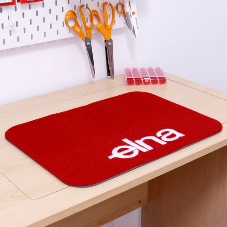 Czerwona podkładka filcowa ELNA o wymiarach 44,5 x 32,5 cm, z białym logo elna. Produkt leży na drewnianym blacie, obok widoczne nożyczki i organizer na akcesoria. Kształt prostokątny z zaokrąglonymi krawędziami.