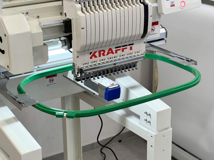 Hafciarka KRAFFT KF-CT1501H, jednogłowicowa, 15-igłowa, z zielonym obrębem do haftu. Widok z boku, z bliska na głowicę maszyny z logo KRAFFT oraz mechanizmem igieł. Tło jasne, stół roboczy w kolorze białym.