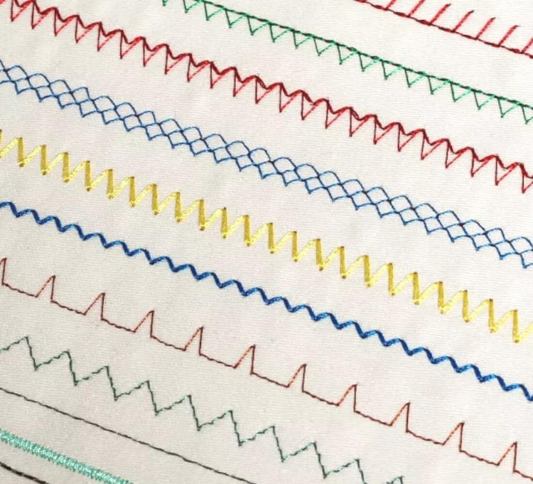 Zbliżenie na różne ściegi wykonane maszyną do szycia Janome JUNO E1015, w tym wzory w kolorach czerwonym, zielonym, niebieskim i żółtym, na jasnym tle. Widoczna różnorodność ściegów dekoracyjnych i użytkowych.