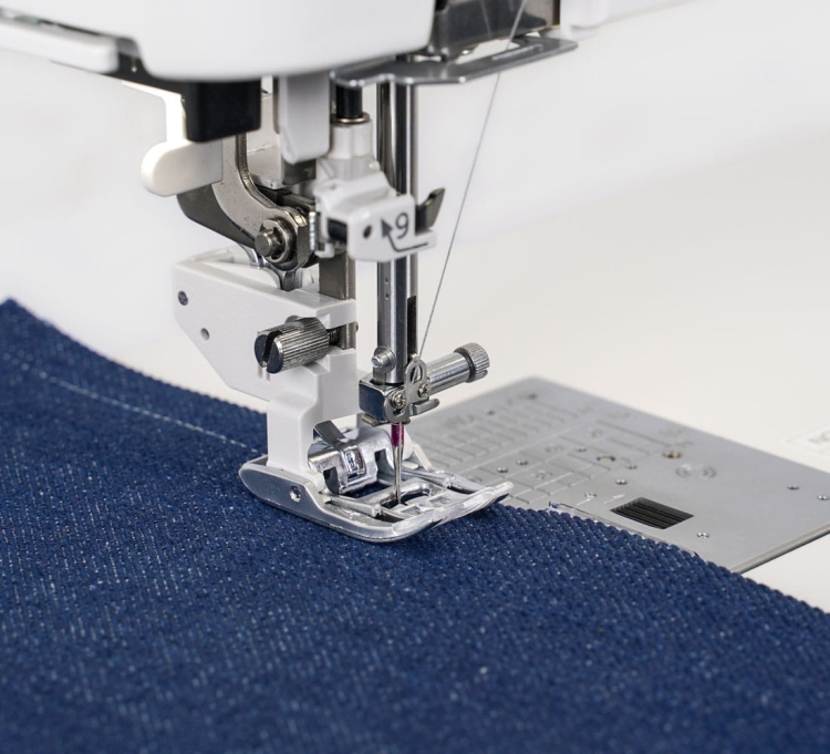 Maszyna do szycia Janome Continental M8 Professional CM8P, widok z bliska na stopkę i igłę, szyjąca ciemnoniebieski materiał jeansowy. Widać elementy regulacyjne oraz płytkę z przyciskami na obudowie. Tło jasne i neutralne.