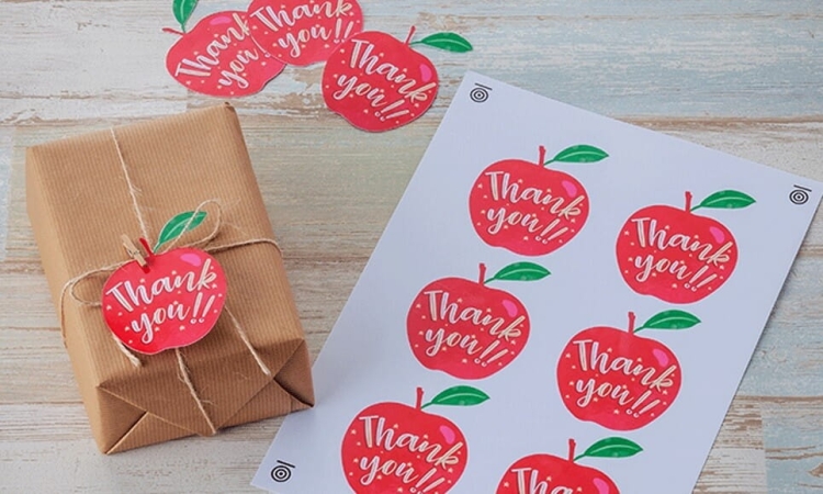 Kolorowy arkusz samoprzylepnych naklejek w kształcie jabłek z napisem Thank you!!. W tle widoczny prezent zapakowany w brązowy papier, ozdobiony naklejką jabłka. Naklejki mają zielone liście i są rozmieszczone w równych odstępach.