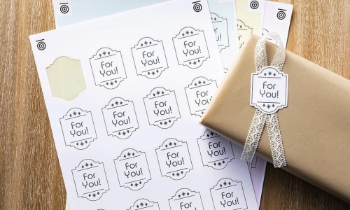 Kod aktywacyjny do funkcji cięcia po znacznikach Print to Cut (CADXPRNTCUT1) przedstawia arkusze z etykietami For You! w różnych kolorach, na tle brązowego opakowania z wstążką. Widoczna jest także przezroczysta naklejka na jednym z arkuszy.